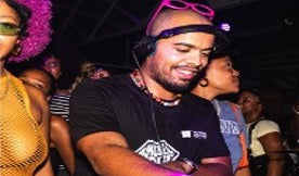 A man at a nightclub or party wearing a black t-shirt, a black cap backwards with pink sunglasses on top, and a necklace, with other people visible in the background and foreground.