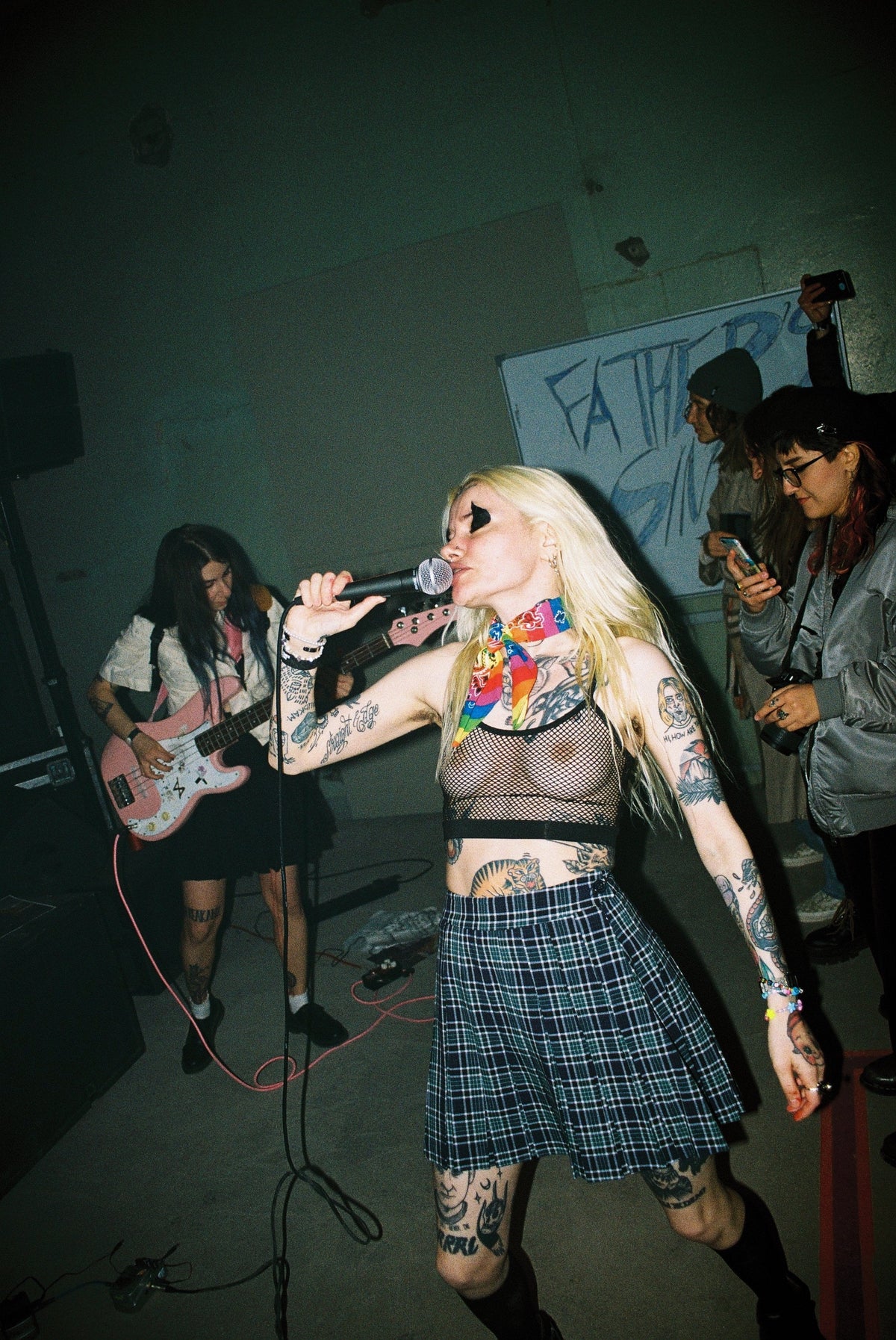 A tattooed blonde woman singing into a microphone on stage, wearing a sheer black crop top, a blue plaid skirt, and a rainbow scarf, with band members and a graffiti-covered wall in the background.
