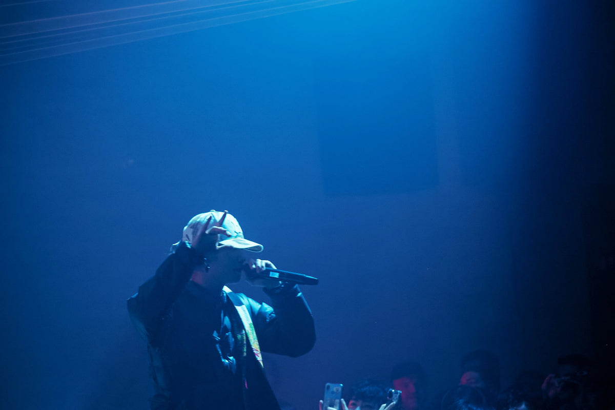 A person on a stage wearing a light-colored baseball cap and dark clothing, holding a microphone under bright blue stage lighting.