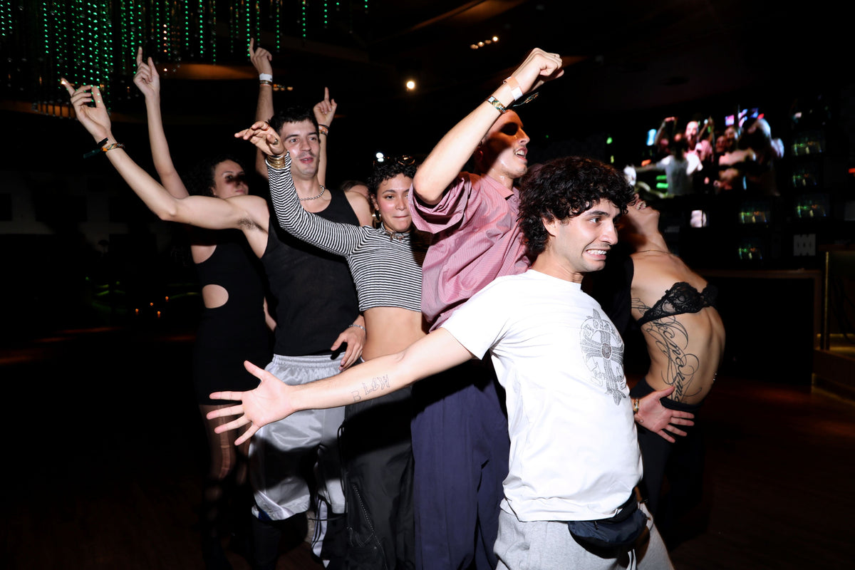 A group of young people dancing energetically in a dimly lit nightclub or bar, with raised arms and a festive atmosphere in the background.