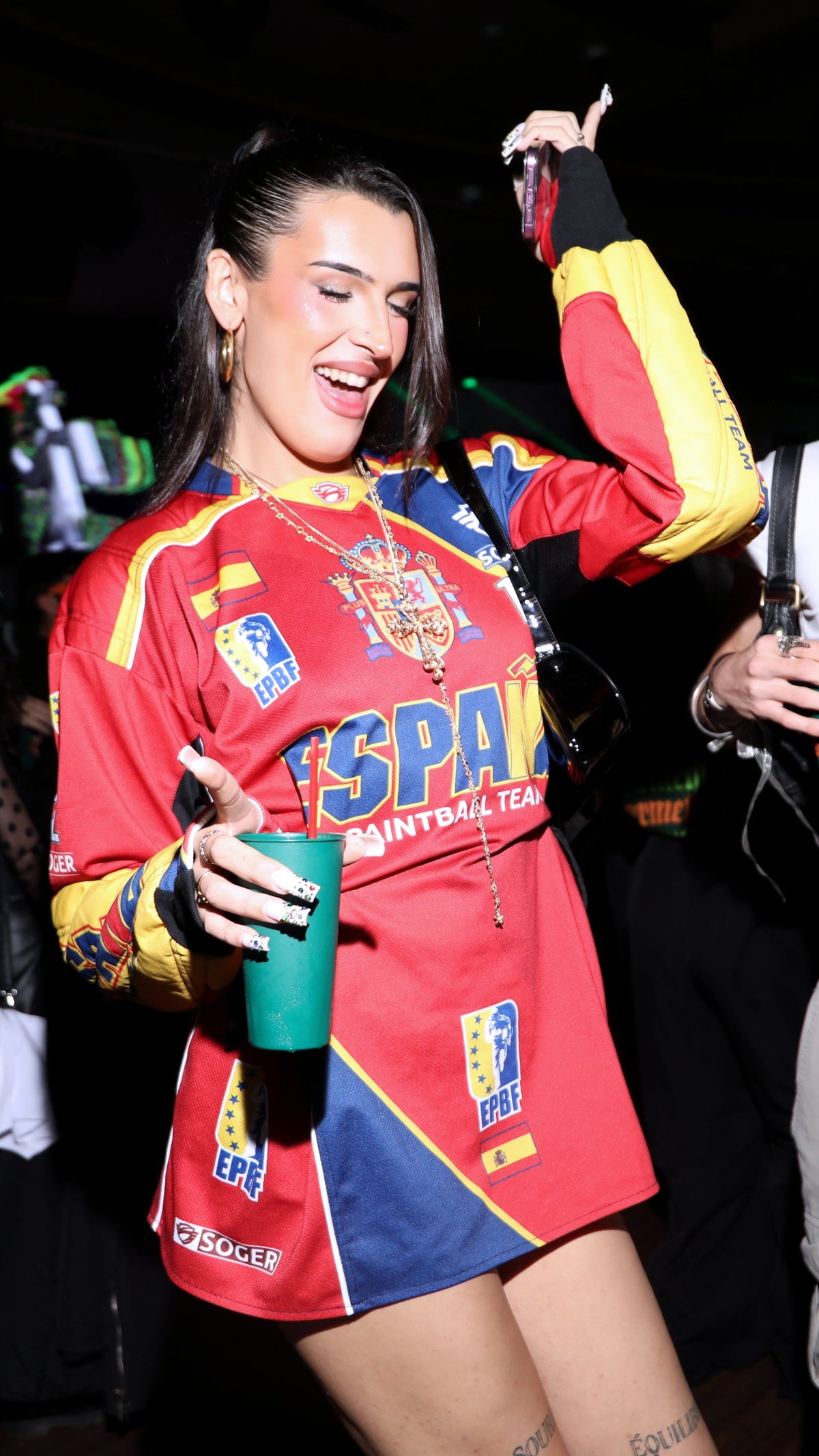 A woman wearing a red, yellow, and blue Spain paintball team long-sleeve jersey dress, holding a green cup in a dark, social setting.
