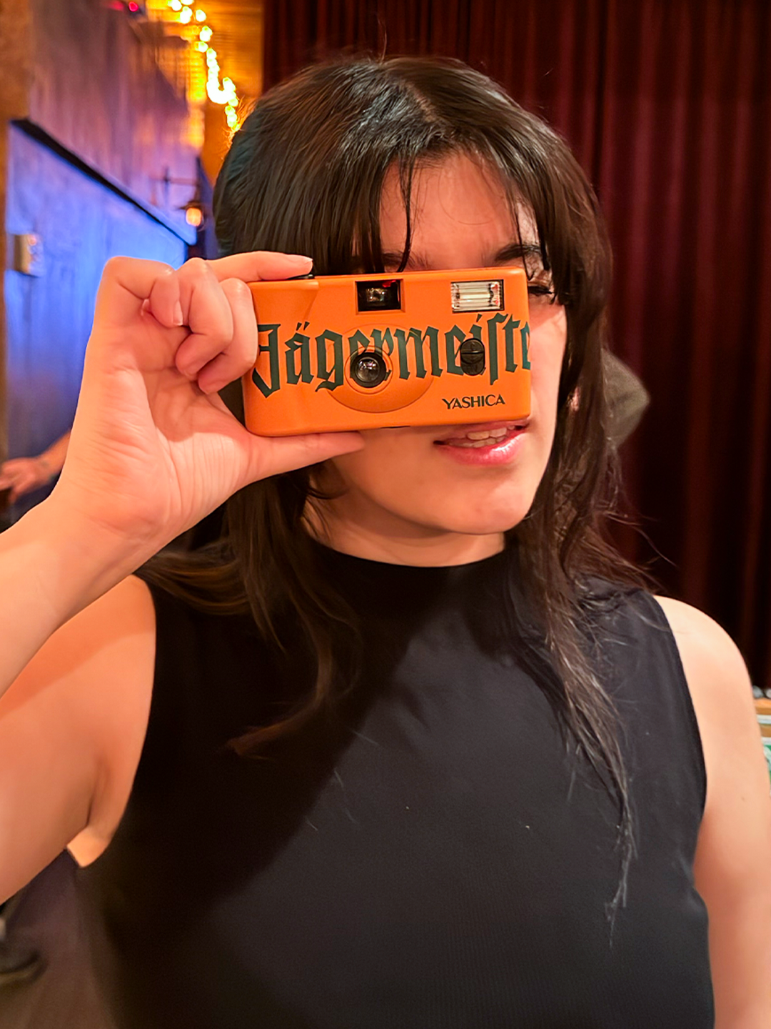 Persona che tiene davanti al viso una fotocamera arancione con marchio 'Jägermeister'.