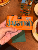 Fotocamera arancione con marchio 'Jägermeister' tenuta da una mano su un tavolo di legno.