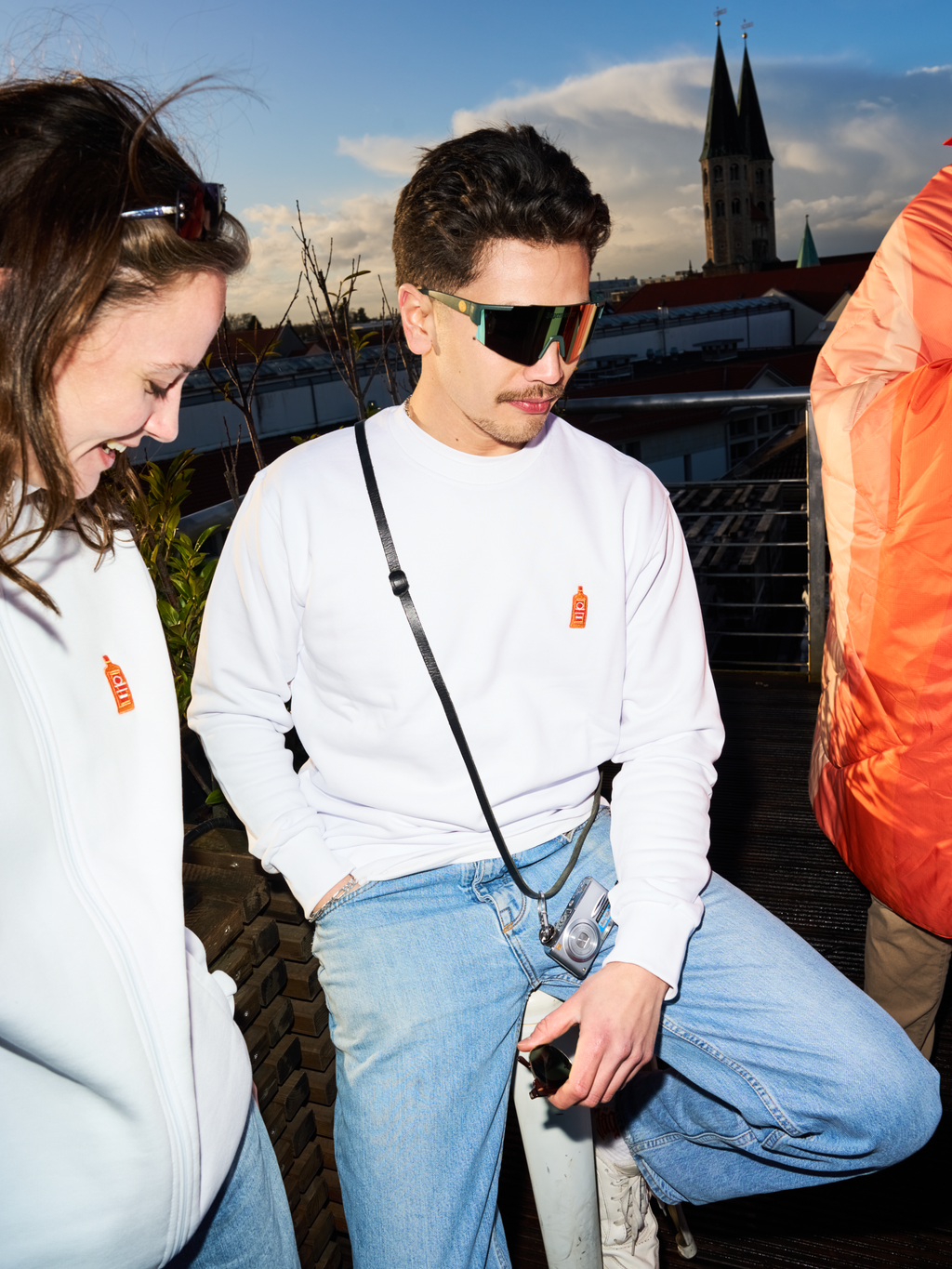Due persone con felpe bianche Jägermeister e occhiali rapidi Jägermeister su una terrazza panoramica, con un campanile e lo skyline della città sullo sfondo.
