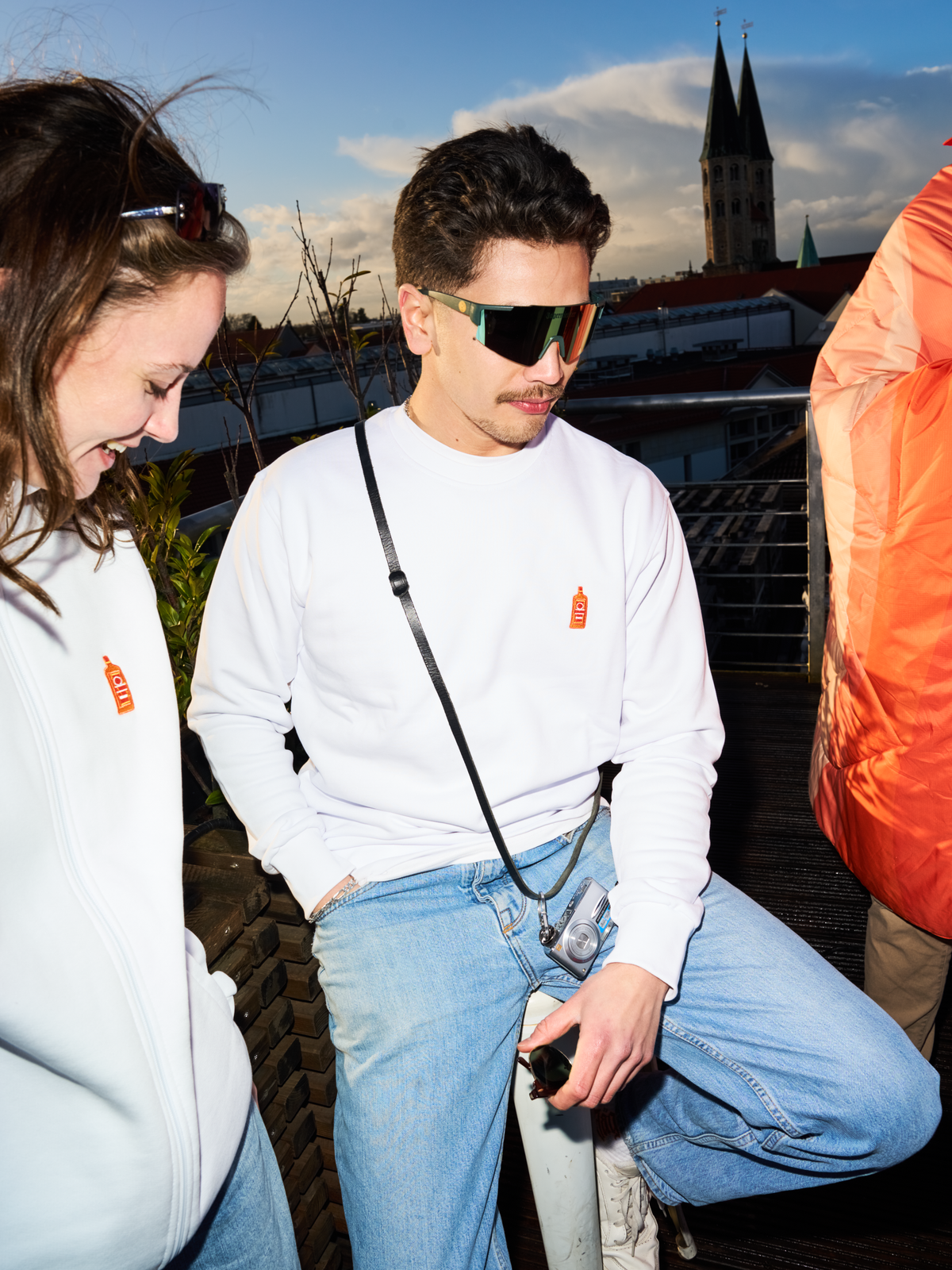 Due persone con felpe bianche Jägermeister e occhiali rapidi Jägermeister su una terrazza panoramica, con un campanile e lo skyline della città sullo sfondo.