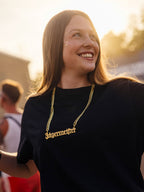 Donna con maglietta nera e collana con catena dorata e charm Jägermeister, sorridente all’aperto in un contesto da festival.
