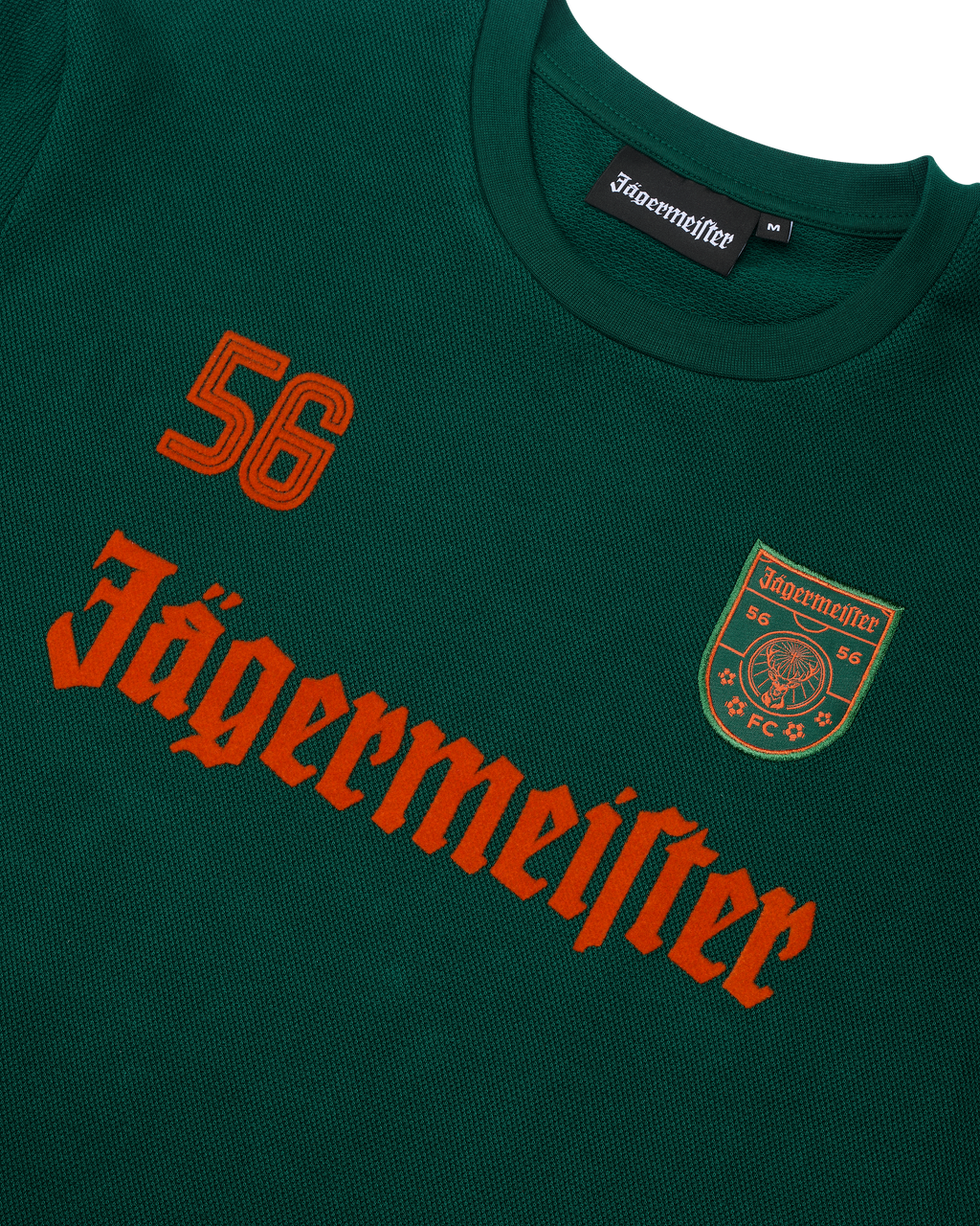 Maglia a maniche lunghe verde Jägermeister con scritta Jägermeister arancione e un '56'.