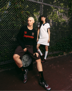 Persona che indossa un abito polo bianco Jägermeister in piedi accanto a una rete metallica, con un’altra persona seduta davanti che indossa un maglione nero Jägermeister e pantaloncini. Ambientazione urbana all’aperto con vegetazione sullo sfondo.