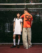 Due modelli in piedi su un campo da tennis rosso davanti a una rete metallica. La modella a sinistra indossa un abito polo bianco Jägermeister con rifiniture verde scuro, calzini bianchi e scarpe nere. Il ragazzo a destra indossa pantaloni beige e una jersey arancione Jägermeister.