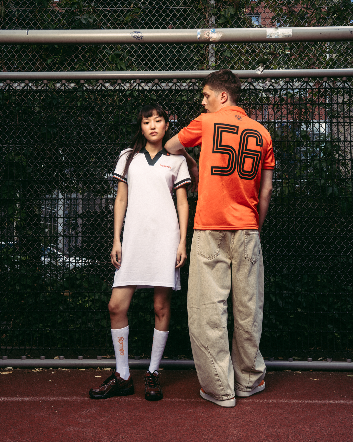 Due modelli in piedi su un campo da tennis rosso davanti a una rete metallica. La modella a sinistra indossa un abito polo bianco Jägermeister con rifiniture verde scuro, calzini bianchi e scarpe nere. Il ragazzo a destra indossa pantaloni beige e una jersey arancione Jägermeister.
