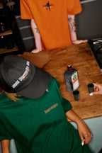Persona che indossa una maglia verde 'Jägermeister' e cappellino nero Jägermeister Racing Cap seduta a un tavolo con prodotti Jägermeister sopra.