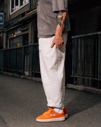 A person standing outdoors on a street wearing loose white trousers, a gray t-shirt, and bright orange low-top sneakers. The photo is cropped from the chest down, with the person's right arm visible and the background showing a dark urban alley or street setting.