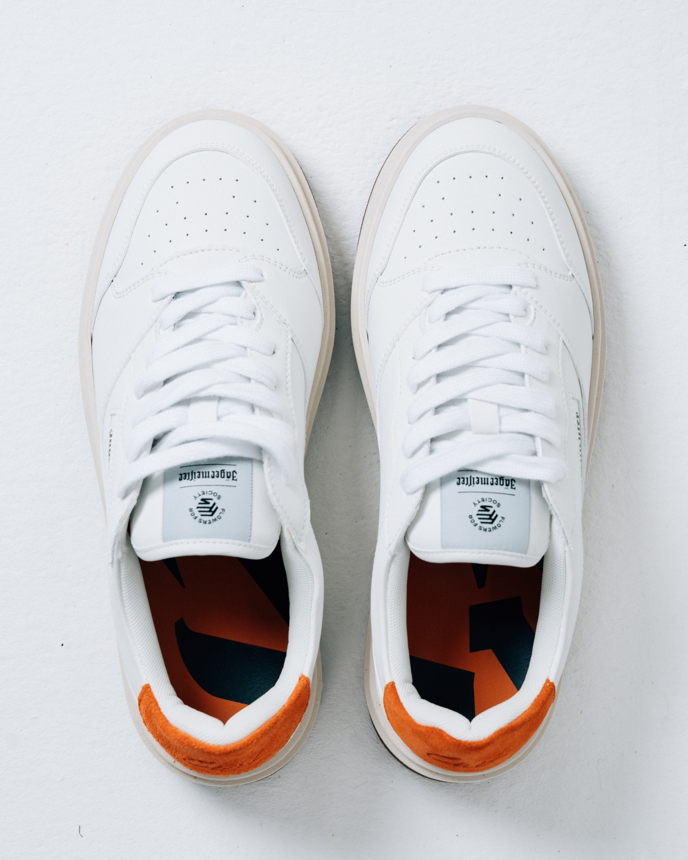 A pair of white Jägermeister sneakers shown from a top-down view on a light gray background. The shoes feature white laces, a white leather upper, and a bright orange heel accent.