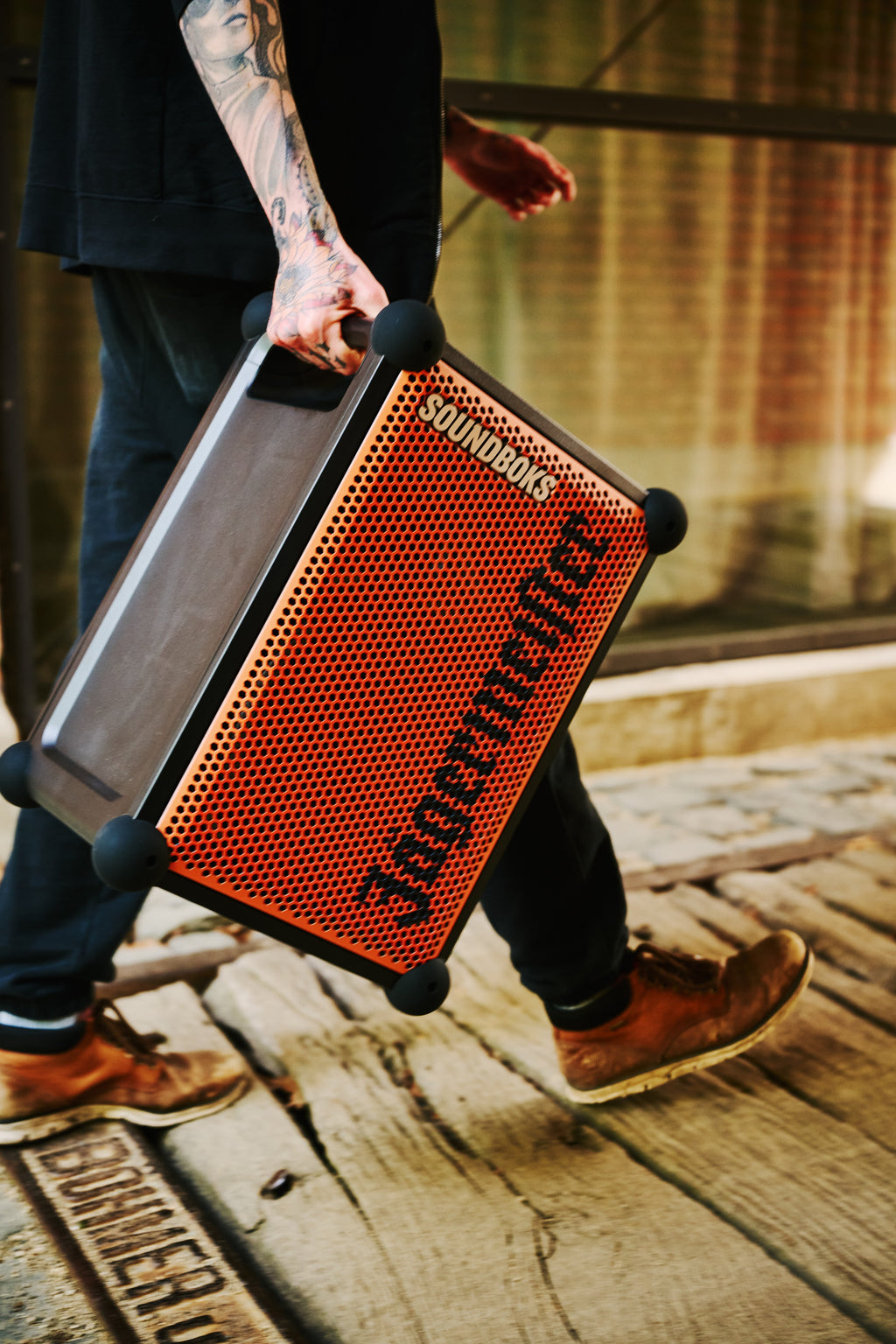 Persona che tiene in mano uno speaker portatile Soundboks arancione e nero con branding 'Jägermeister' mentre cammina su un pavimento in legno