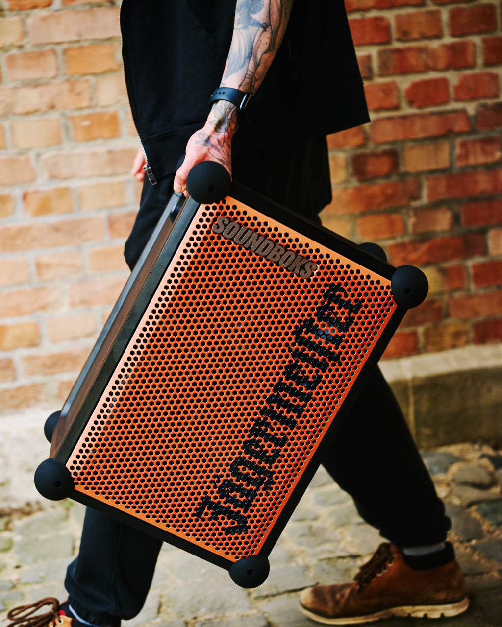 Persona che tiene in mano uno speaker portatile Soundboks arancione e nero con branding 'Jägermeister' davanti a una parete di mattoni.