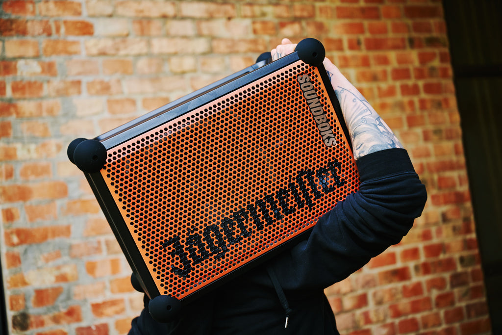 Persona che tiene in mano uno speaker portatile Soundboks arancione e nero con branding 'Jägermeister' davanti a una parete di mattoni.