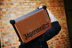 Persona che tiene in mano uno speaker portatile Soundboks arancione e nero con branding 'Jägermeister' davanti a una parete di mattoni.