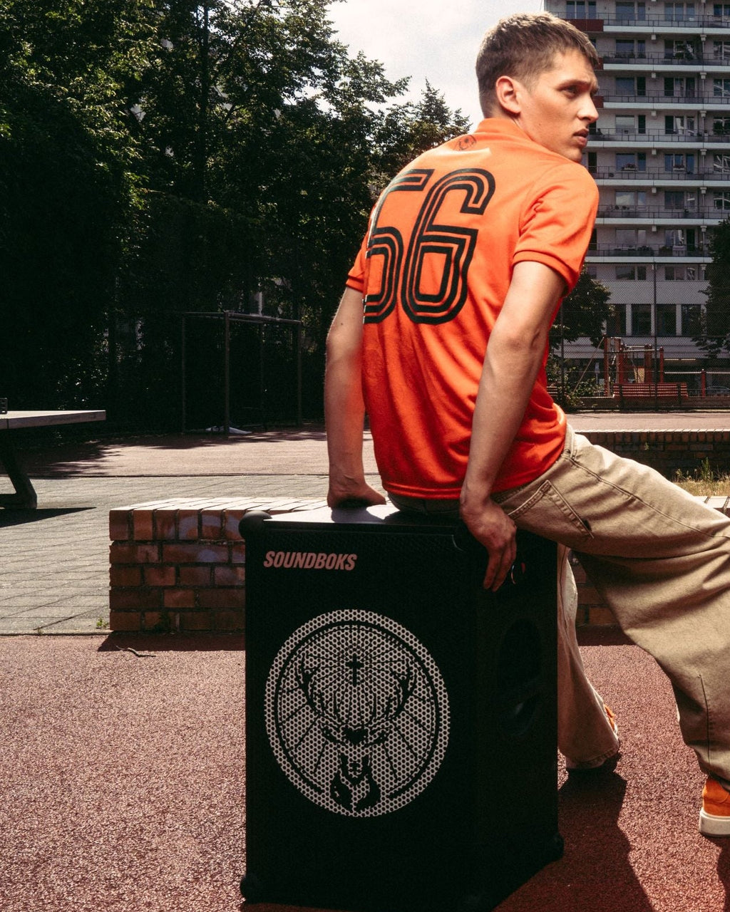 Un uomo in una maglia arancione Jägermeister e pantaloni beige è seduto su uno speaker Soundboks nero con il logo del marchio Jägermeister