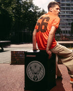 Un uomo in una maglia arancione Jägermeister e pantaloni beige è seduto su uno speaker Soundboks nero con il logo del marchio Jägermeister