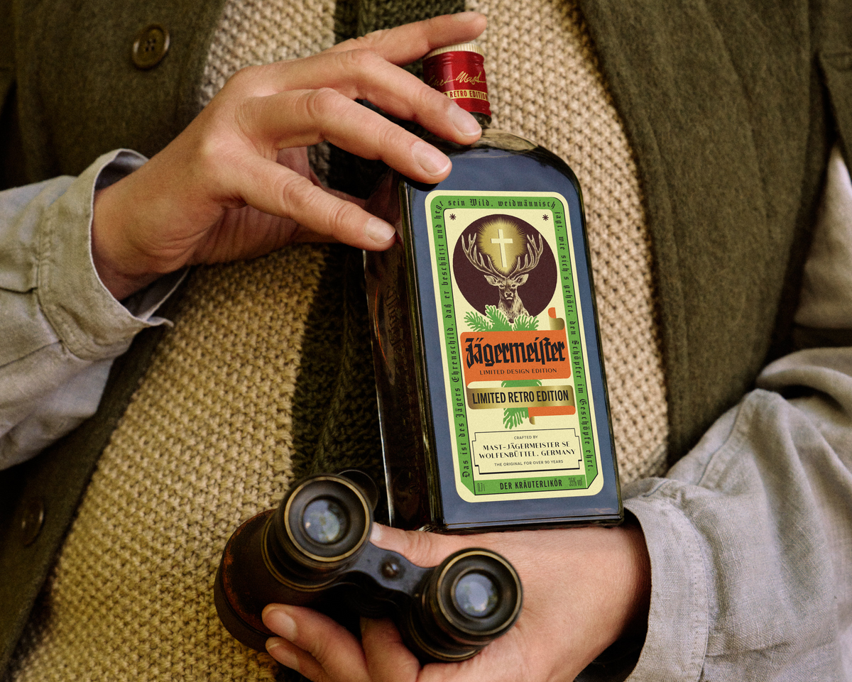 A person holds a retro Jägermeister bottle, while also holding a pair of binoculars. The person is wearing a textured beige sweater and an olive green jacket.