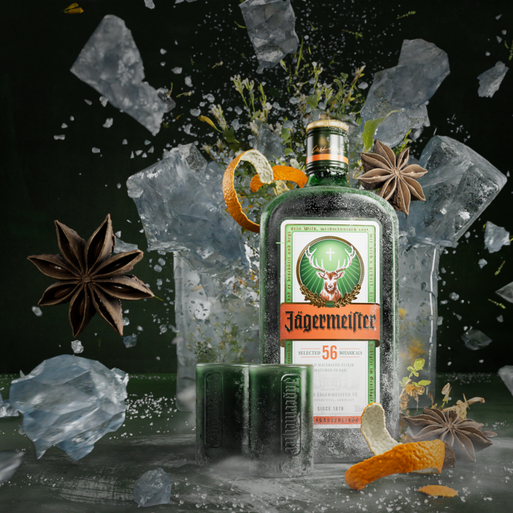 A bottle of Jägermeister, two green shot glasses, and large ice chunks are featured in a dynamic, high-speed photograph. Anise stars, orange peel, and herbs are suspended in the air.