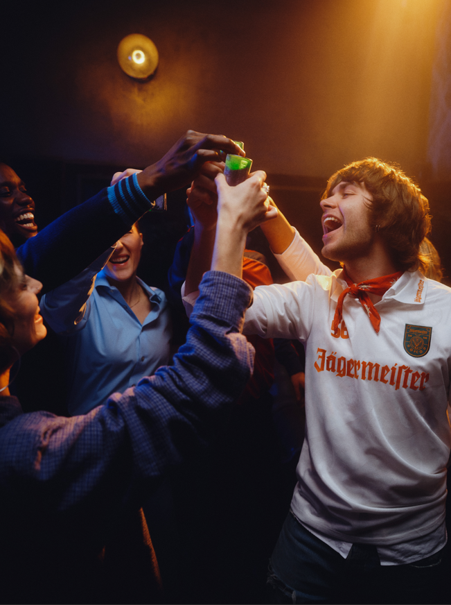 Gruppo di persone che festeggia con una persona che indossa una maglietta Jägermeister in una stanza poco illuminata.